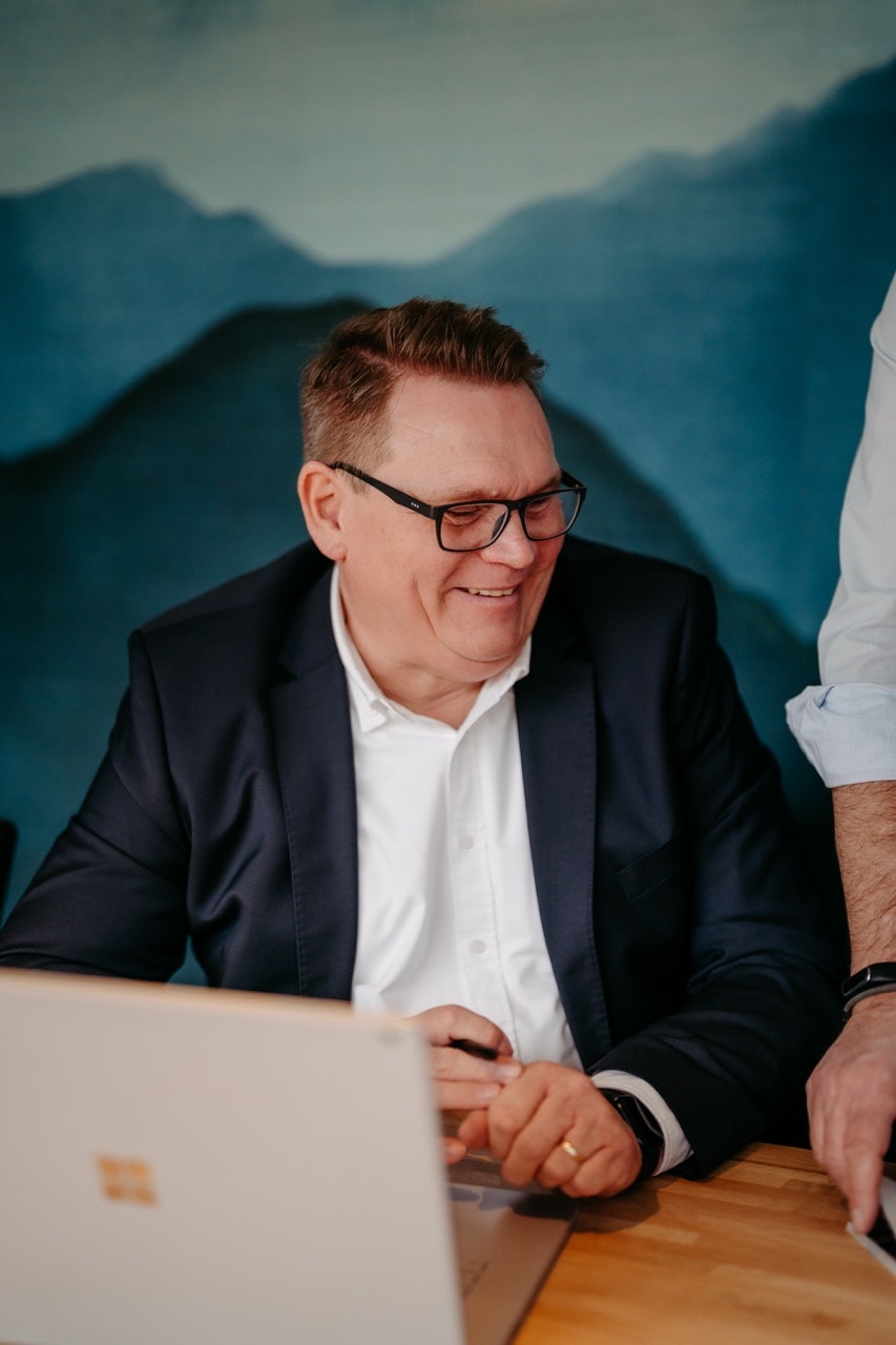 Alwin Justen lachend am Laptop vor einer Bergkulisse-Tapete